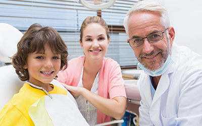 young boy sitting in dentist chair with mother and dentist all smiling together